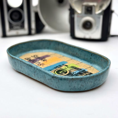 Handmade ceramic catchall tray with a teal glazed rim and a retro camera illustration reading “Smile, darling” on a speckled stoneware base.