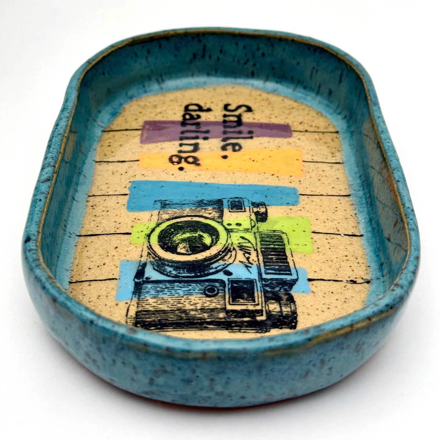 Handmade ceramic catchall tray with a teal glazed rim and a retro camera illustration reading “Smile, darling” on a speckled stoneware base.