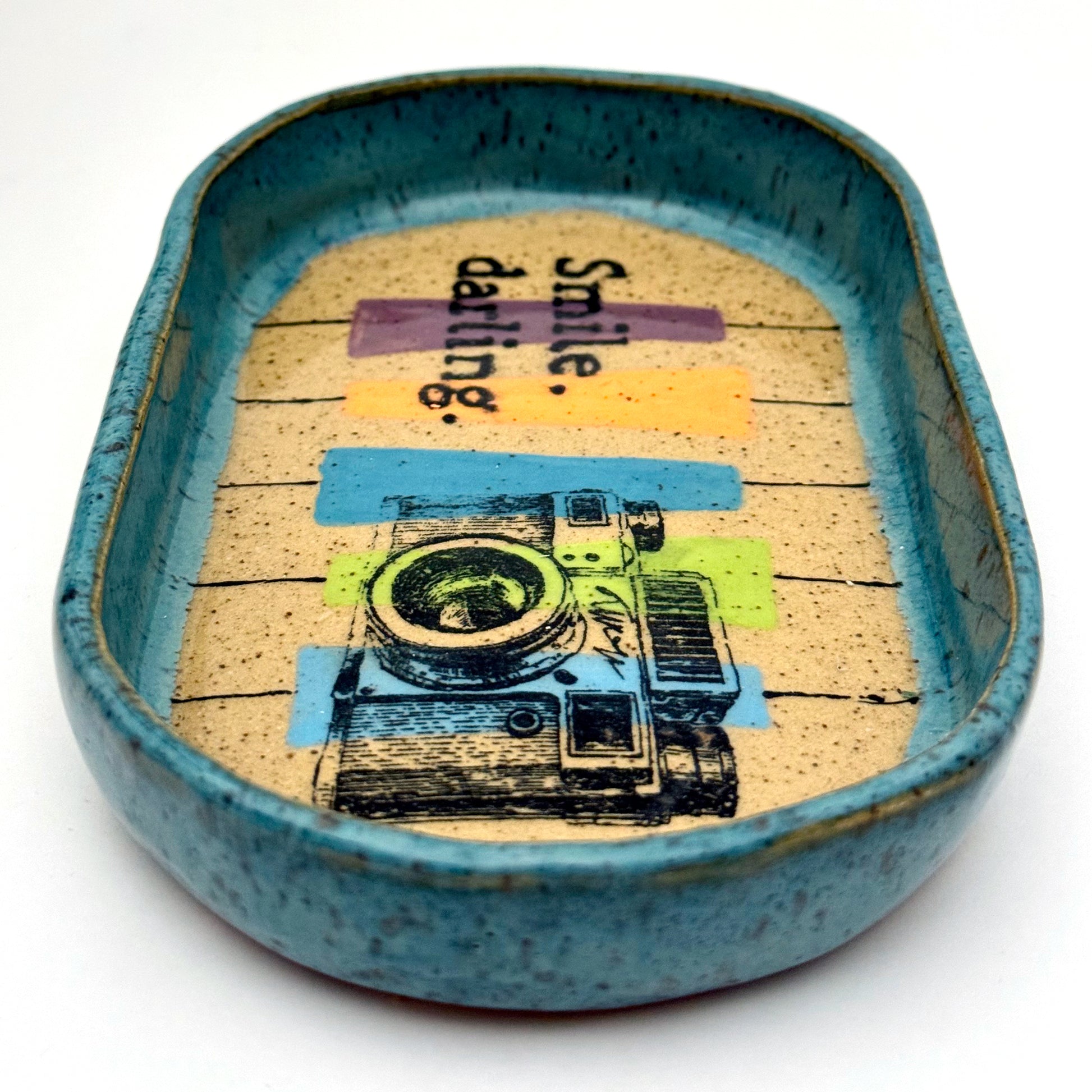 Handmade ceramic catchall tray with a teal glazed rim and a retro camera illustration reading “Smile, darling” on a speckled stoneware base.
