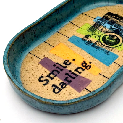 Handmade ceramic catchall tray with a teal glazed rim and a retro camera illustration reading “Smile, darling” on a speckled stoneware base.
