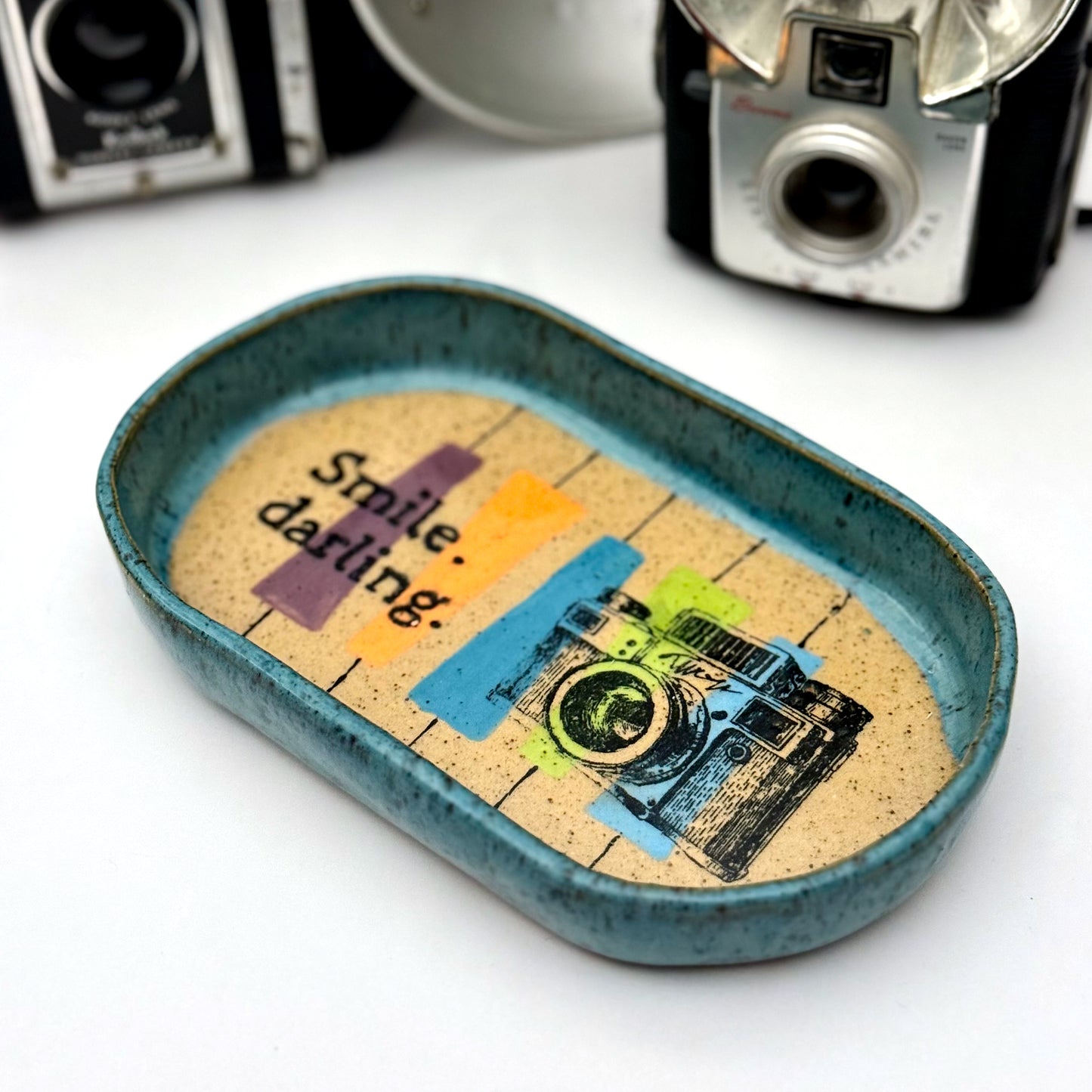 Handmade ceramic catchall tray with a teal glazed rim and a retro camera illustration reading “Smile, darling” on a speckled stoneware base.