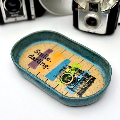 Handmade ceramic catchall tray with a teal glazed rim and a retro camera illustration reading “Smile, darling” on a speckled stoneware base.