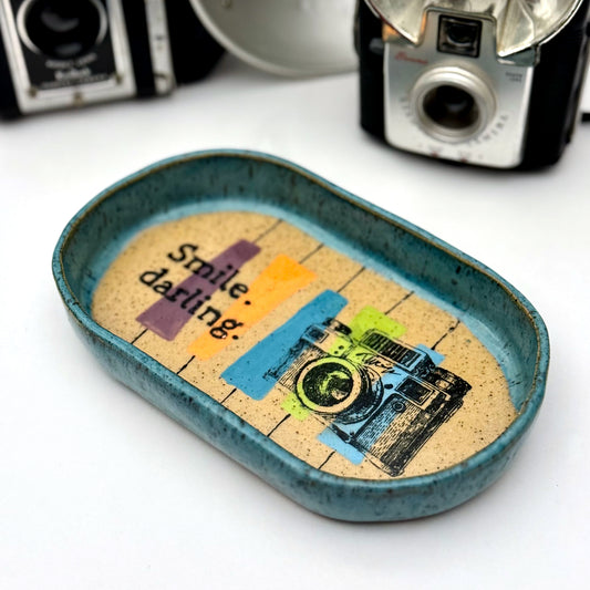 Handmade ceramic catchall tray with a teal glazed rim and a retro camera illustration reading “Smile, darling” on a speckled stoneware base.