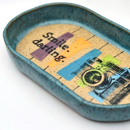 Handmade ceramic catchall tray with a teal glazed rim and a retro camera illustration reading “Smile, darling” on a speckled stoneware base.