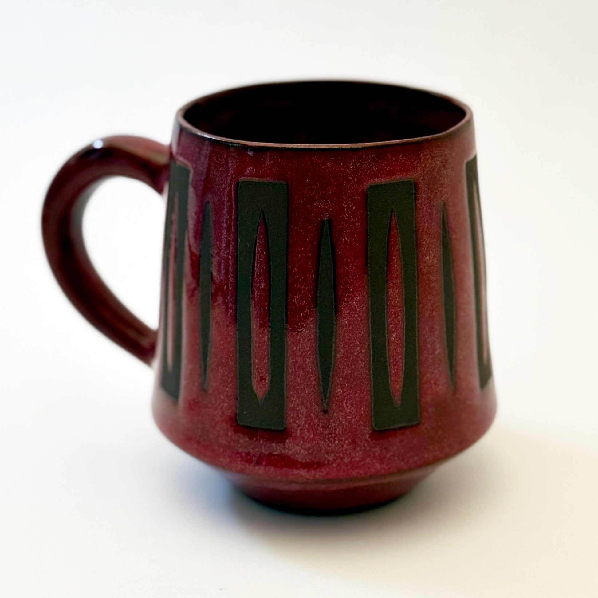 Hand-thrown stoneware mug with a deep burgundy glaze and repeating elongated geometric pattern in dark teal-black. The bold mid-century-inspired design wraps around the slightly tapered mug, which features a curved handle and glossy finish. Photographed against a soft white background.