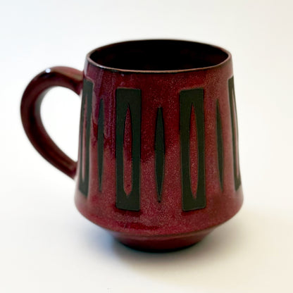Hand-thrown stoneware mug with a deep burgundy glaze and repeating elongated geometric pattern in dark teal-black. The bold mid-century-inspired design wraps around the slightly tapered mug, which features a curved handle and glossy finish. Photographed against a soft white background.
