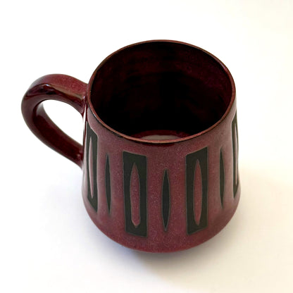 Hand-thrown stoneware mug with a deep burgundy glaze and repeating elongated geometric pattern in dark teal-black. The bold mid-century-inspired design wraps around the slightly tapered mug, which features a curved handle and glossy finish. Photographed against a soft white background.