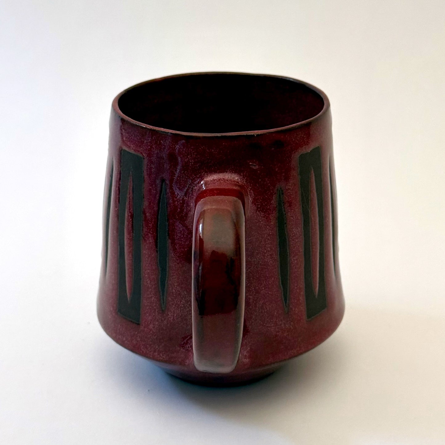 Hand-thrown stoneware mug with a deep burgundy glaze and repeating elongated geometric pattern in dark teal-black. The bold mid-century-inspired design wraps around the slightly tapered mug, which features a curved handle and glossy finish. Photographed against a soft white background.