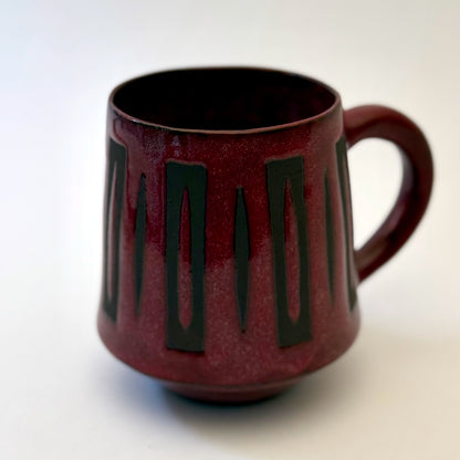 Hand-thrown stoneware mug with a deep burgundy glaze and repeating elongated geometric pattern in dark teal-black. The bold mid-century-inspired design wraps around the slightly tapered mug, which features a curved handle and glossy finish. Photographed against a soft white background.