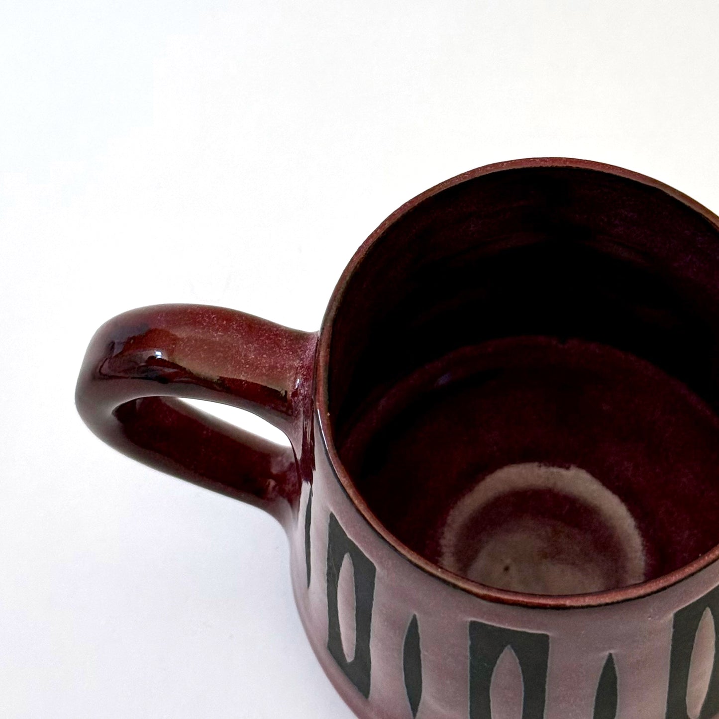 Hand-thrown stoneware mug with a deep burgundy glaze and repeating elongated geometric pattern in dark teal-black. The bold mid-century-inspired design wraps around the slightly tapered mug, which features a curved handle and glossy finish. Photographed against a soft white background.