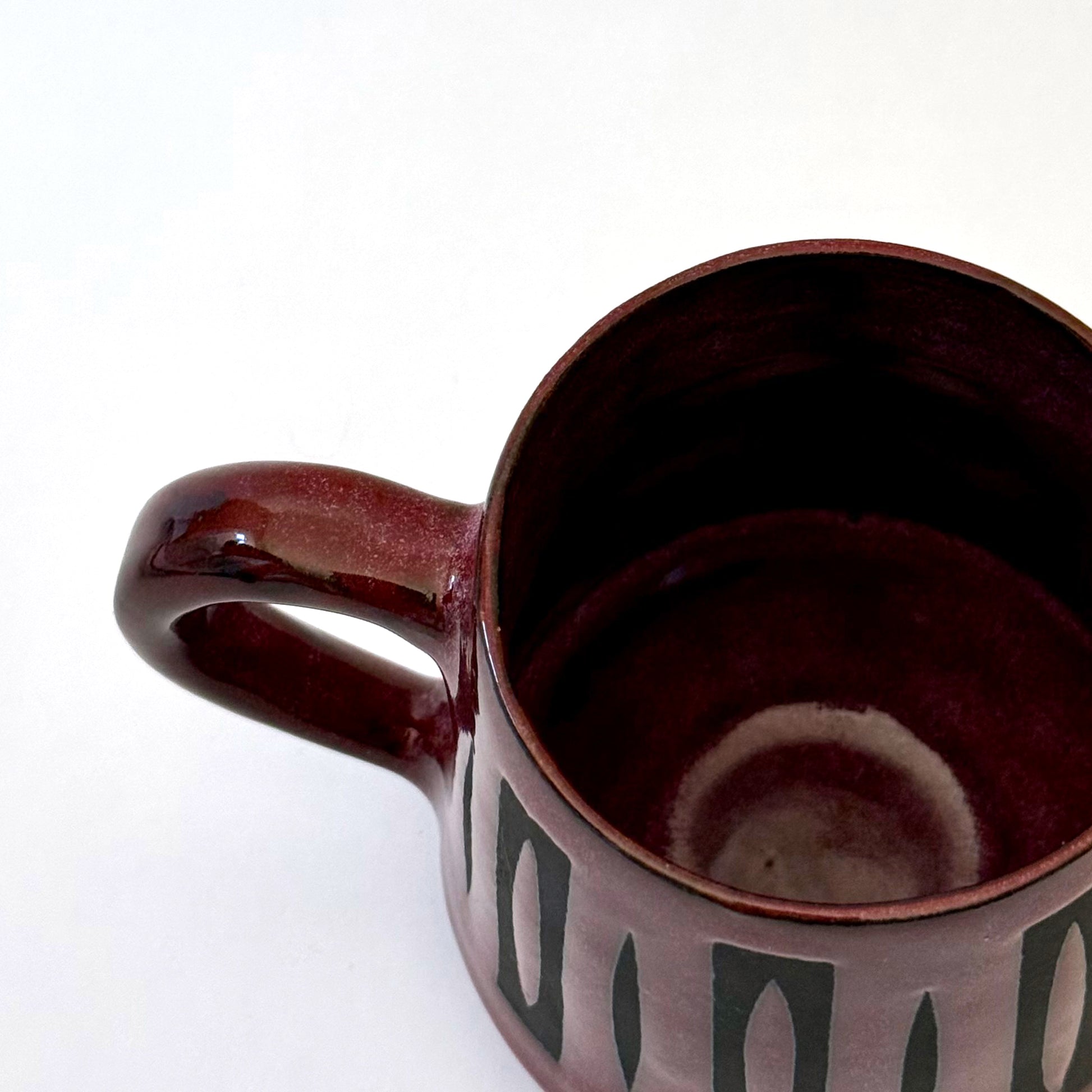 Hand-thrown stoneware mug with a deep burgundy glaze and repeating elongated geometric pattern in dark teal-black. The bold mid-century-inspired design wraps around the slightly tapered mug, which features a curved handle and glossy finish. Photographed against a soft white background.