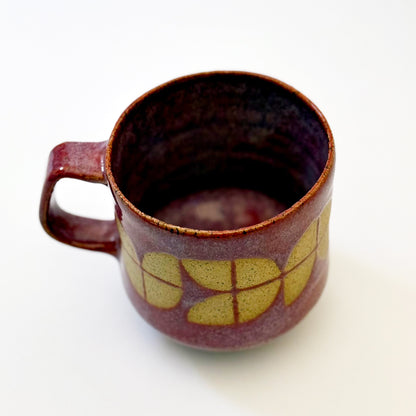 A sturdy, red-glazed mug featuring a repeating geometric tulip pattern in raw, speckled tan stoneware. The design uses sharp lines and a bold, angular handle to create a retro, architectural silhouette.