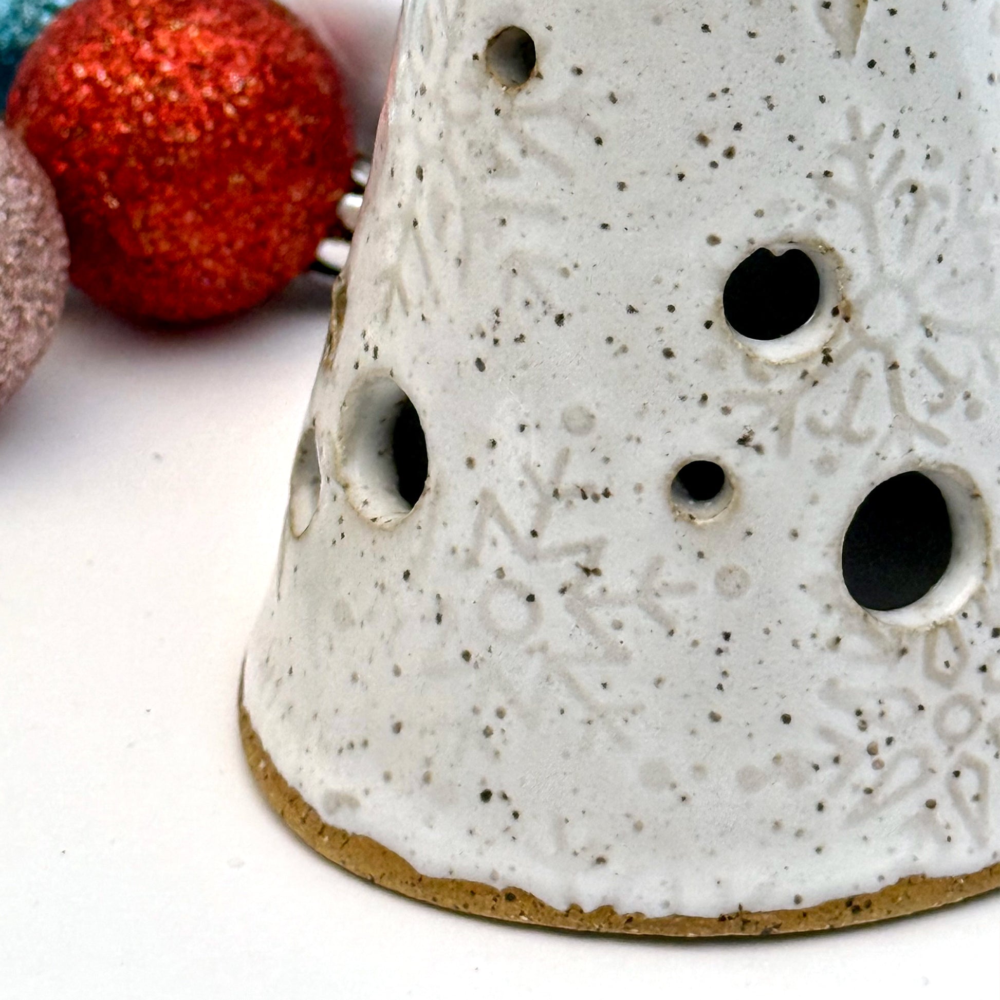 Close-up of a white ceramic tree luminary with embossed snowflake texture and round cutout holes, surrounded by colorful holiday ornaments.