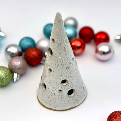 A white ceramic tree luminary with embossed snowflake texture and round cutout holes, surrounded by colorful holiday ornaments.