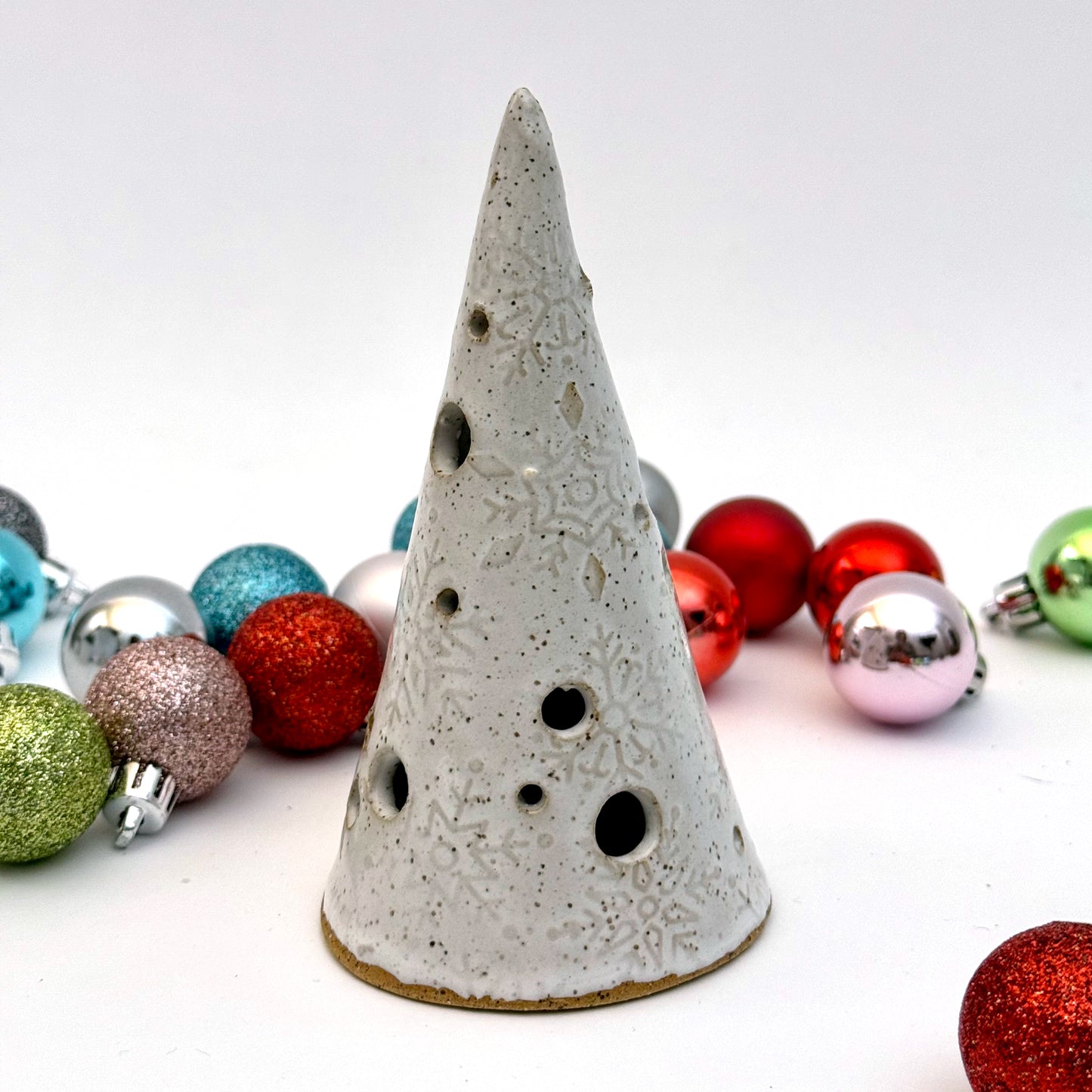 A white ceramic tree luminary with embossed snowflake texture and round cutout holes, surrounded by colorful holiday ornaments.