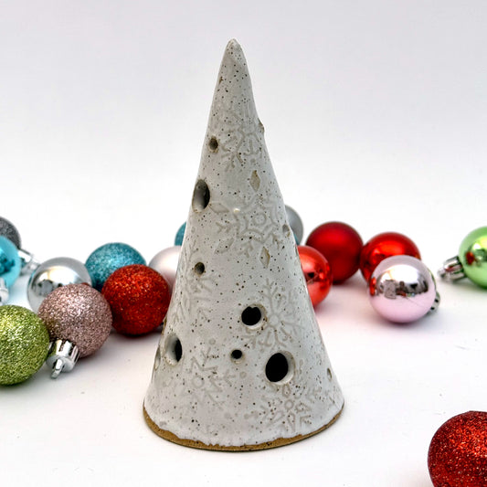 A white ceramic tree luminary with embossed snowflake texture and round cutout holes, surrounded by colorful holiday ornaments.