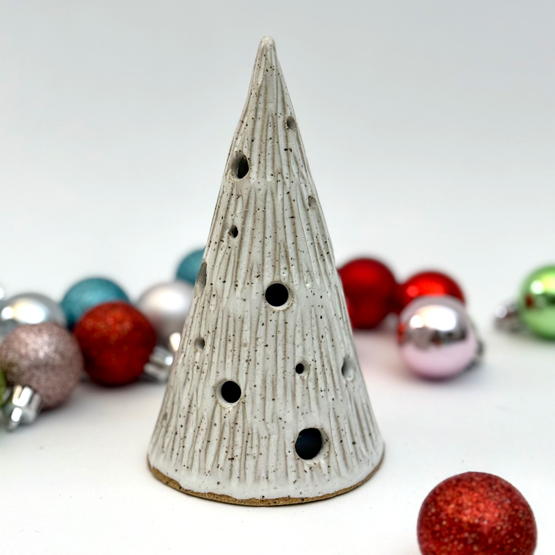 A ceramic tree luminary in a snowy white glaze with carved lines and round holes for light, shown among Christmas ornaments.