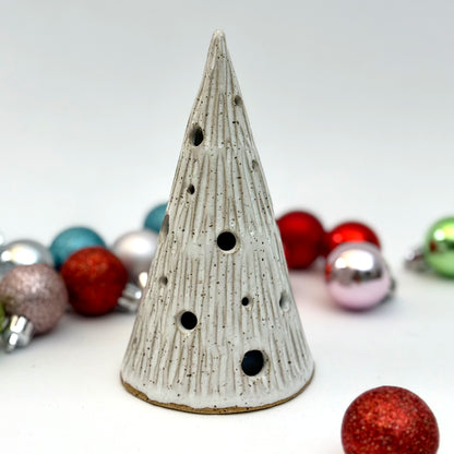 A ceramic tree luminary in a snowy white glaze with carved lines and round holes for light, shown among Christmas ornaments.