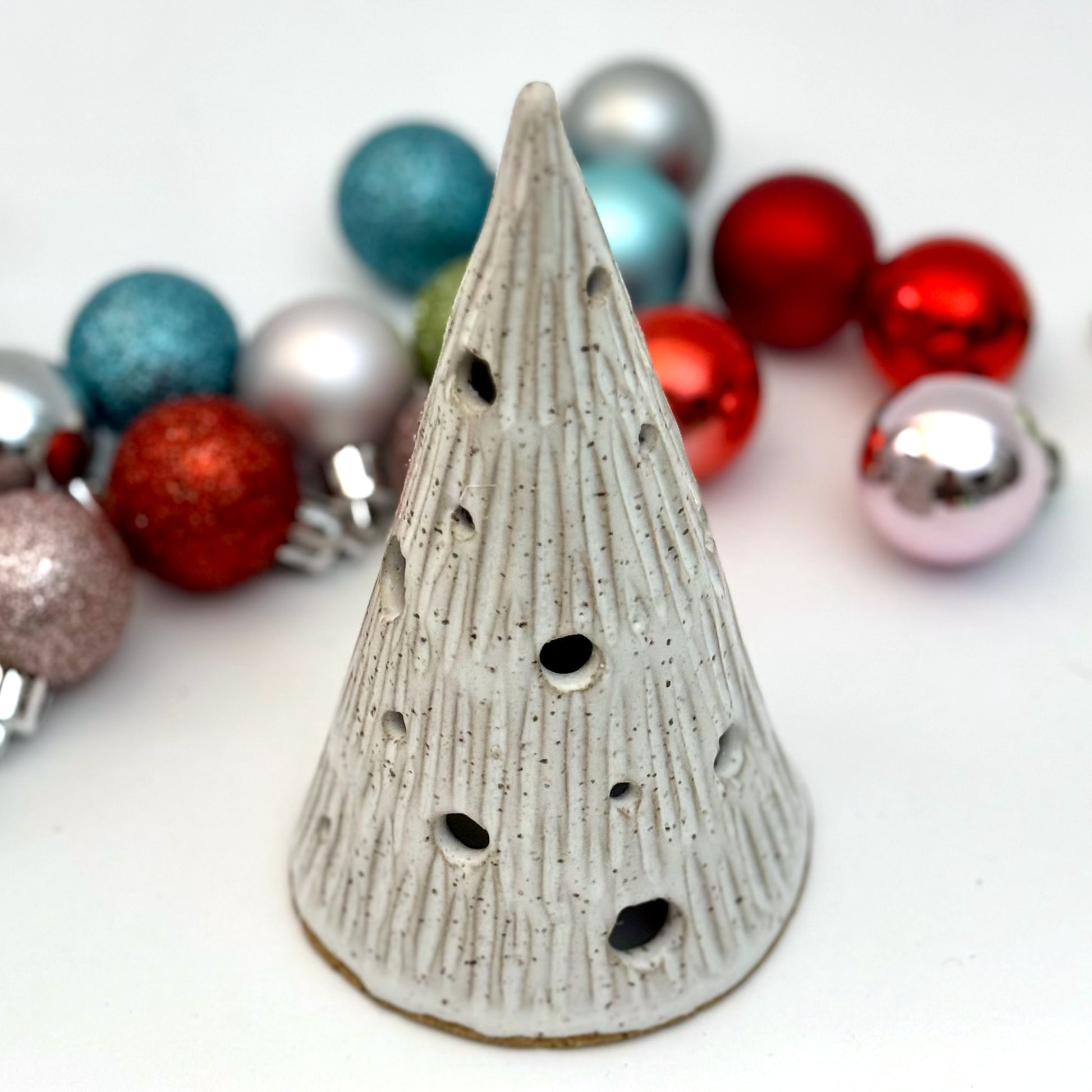 A ceramic tree luminary in a snowy white glaze with carved lines and round holes for light, shown among Christmas ornaments.