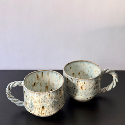 Two handmade ceramic mugs with rounded forms, soft mottled blue glaze, and chunky twisted handles. Each mug has a speckled stoneware surface and an organic, sculptural shape with subtle mid-century character.