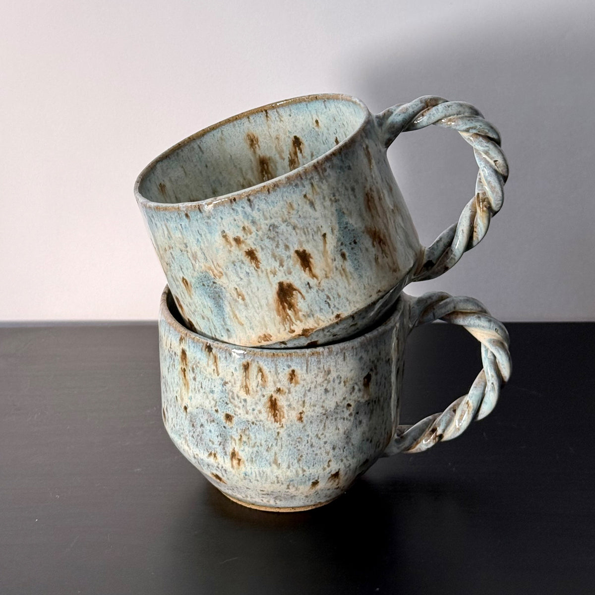 Two handmade ceramic mugs with rounded forms, soft mottled blue glaze, and chunky twisted handles. Each mug has a speckled stoneware surface and an organic, sculptural shape with subtle mid-century character.
