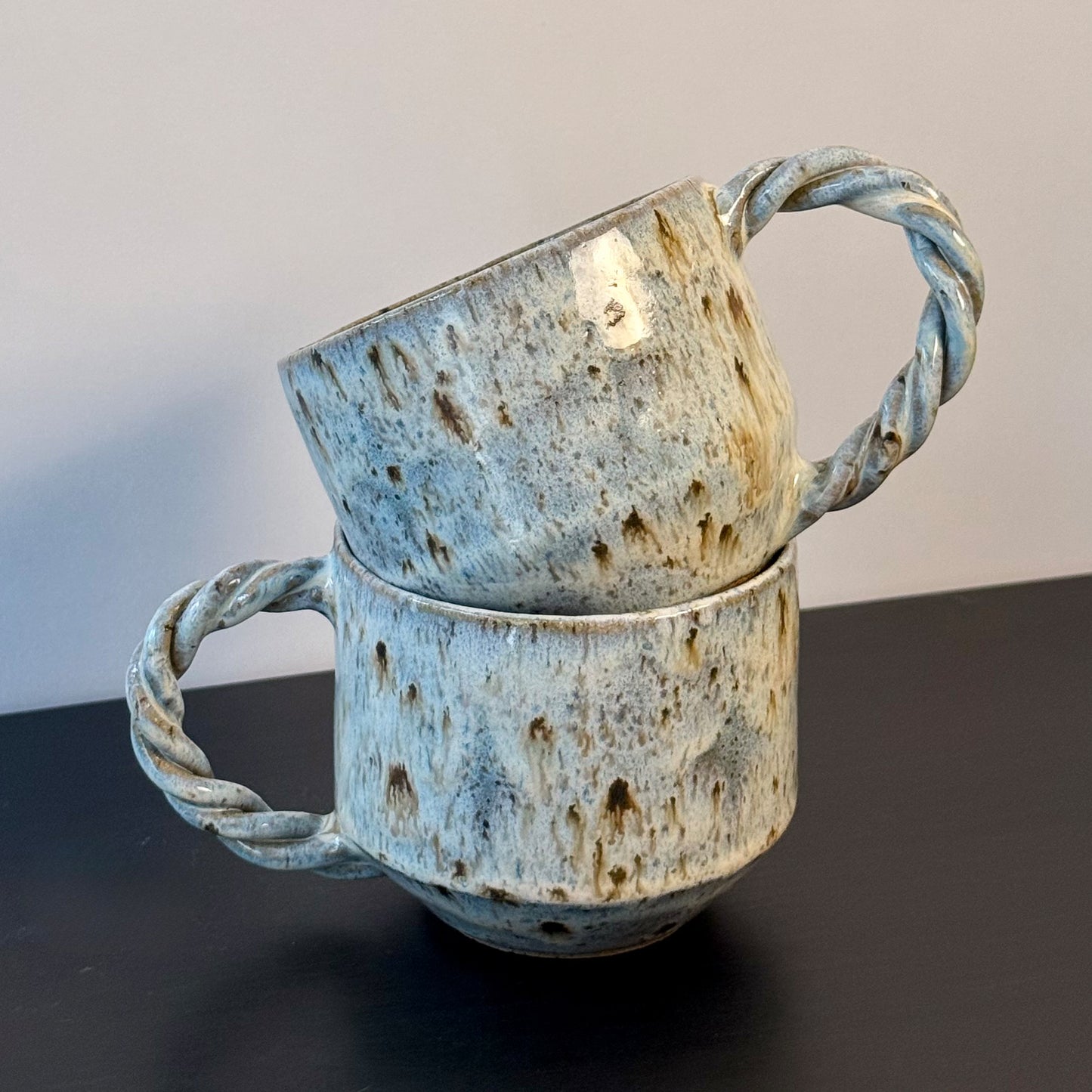 Two handmade ceramic mugs with rounded forms, soft mottled blue glaze, and chunky twisted handles. Each mug has a speckled stoneware surface and an organic, sculptural shape with subtle mid-century character.