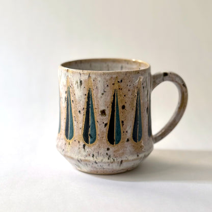 Handmade stoneware mug with a teal-and-black teardrop pattern on a speckled cream glaze, photographed against a white background.