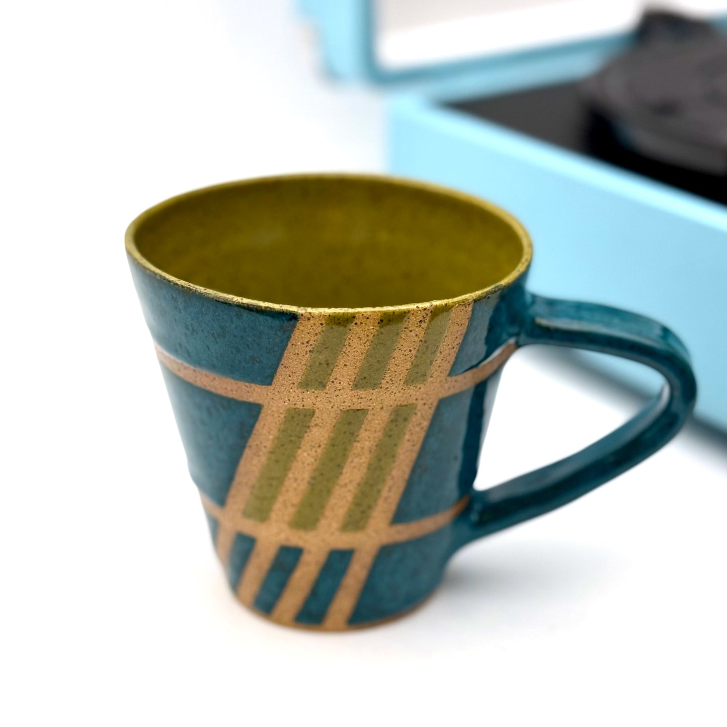 Handmade ceramic mug finished in deep teal and olive glazes with a mid-century–inspired intersecting pattern.