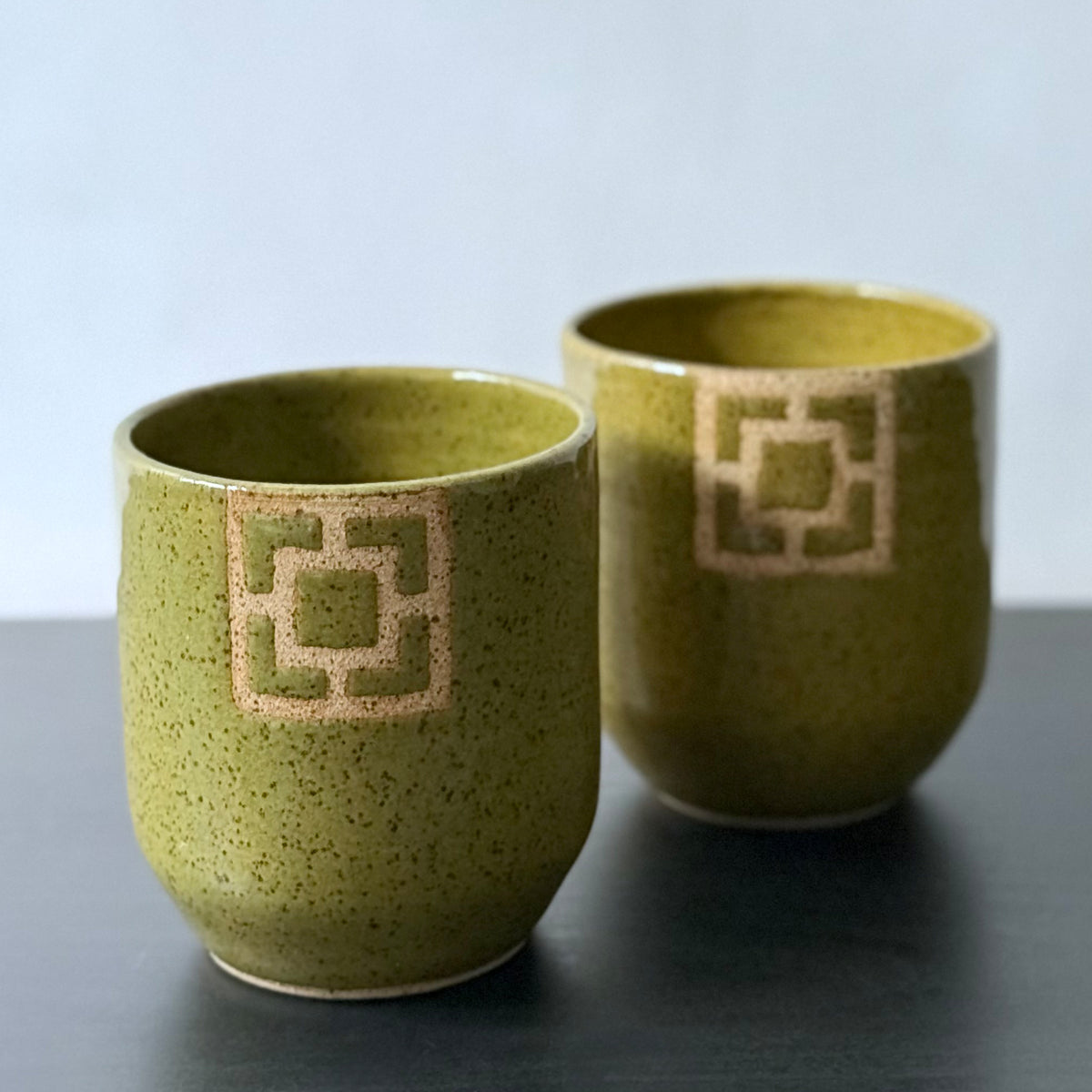 Two handmade ceramic tumblers with an olive-green speckled glaze and rounded shape. Each features a square geometric breeze-block motif on the front, revealing the natural clay and adding a bold mid-century architectural detail.