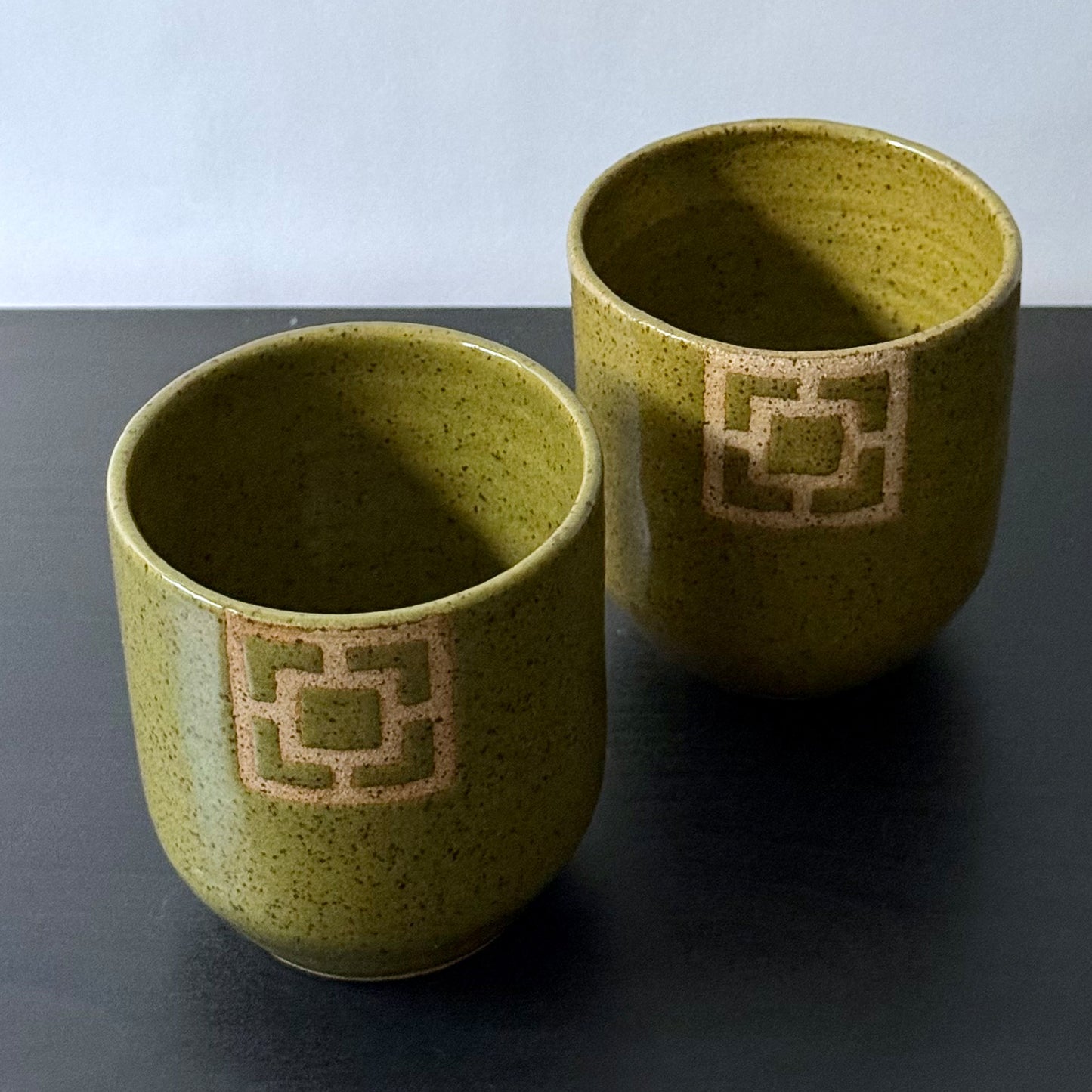 Two handmade ceramic tumblers with an olive-green speckled glaze and rounded shape. Each features a square geometric breeze-block motif on the front, revealing the natural clay and adding a bold mid-century architectural detail.