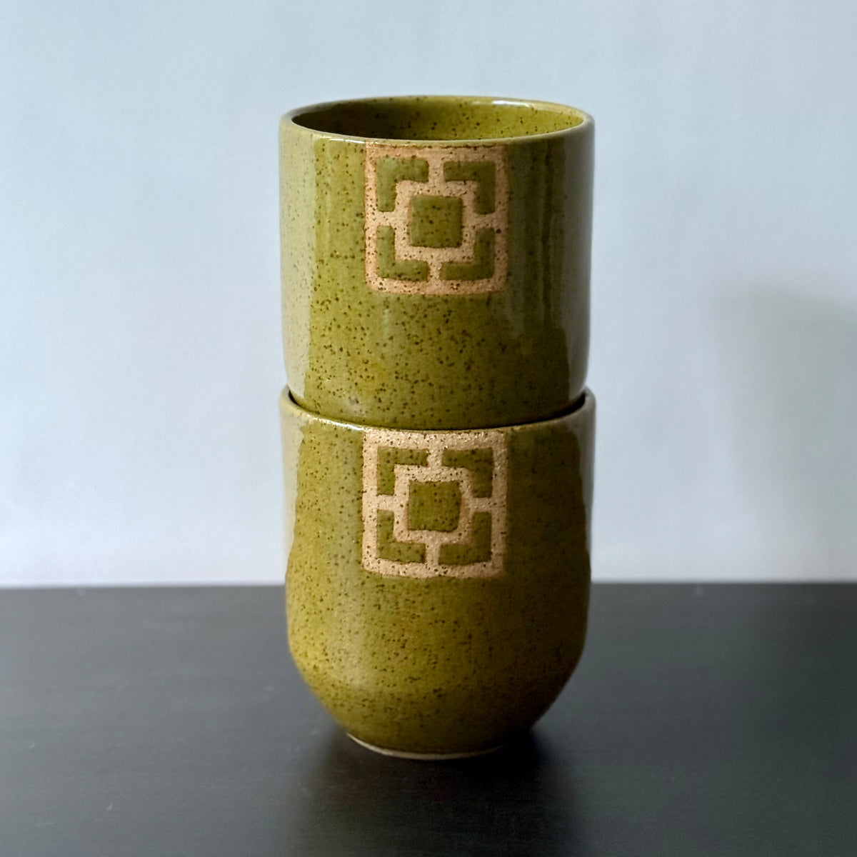Two handmade ceramic tumblers with an olive-green speckled glaze and rounded shape. Each features a square geometric breeze-block motif on the front, revealing the natural clay and adding a bold mid-century architectural detail.