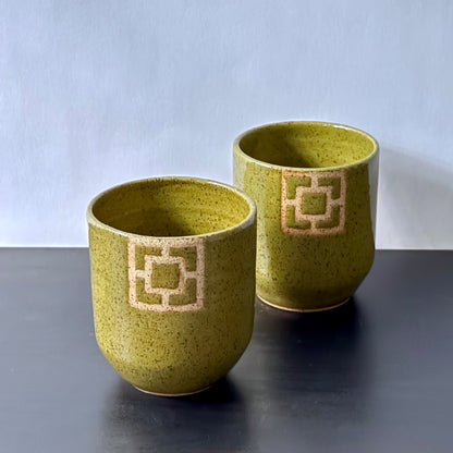 Two handmade ceramic tumblers with an olive-green speckled glaze and rounded shape. Each features a square geometric breeze-block motif on the front, revealing the natural clay and adding a bold mid-century architectural detail.