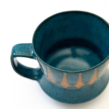 Hand-thrown stoneware mug with layered cobalt and teal blue glaze and a repeating retro geometric pattern in exposed speckled clay. The sculpted mug has a curved waist, angular handle, glossy finish, and mid-century-inspired design. Photographed against a soft white background.