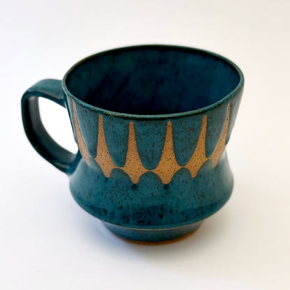 Hand-thrown stoneware mug with layered cobalt and teal blue glaze and a repeating retro geometric pattern in exposed speckled clay. The sculpted mug has a curved waist, angular handle, glossy finish, and mid-century-inspired design. Photographed against a soft white background.