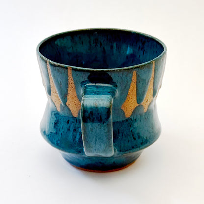 Hand-thrown stoneware mug with layered cobalt and teal blue glaze and a repeating retro geometric pattern in exposed speckled clay. The sculpted mug has a curved waist, angular handle, glossy finish, and mid-century-inspired design. Photographed against a soft white background.