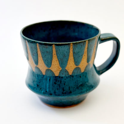Hand-thrown stoneware mug with layered cobalt and teal blue glaze and a repeating retro geometric pattern in exposed speckled clay. The sculpted mug has a curved waist, angular handle, glossy finish, and mid-century-inspired design. Photographed against a soft white background.