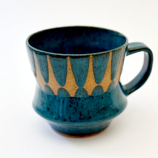 Hand-thrown stoneware mug with layered cobalt and teal blue glaze and a repeating retro geometric pattern in exposed speckled clay. The sculpted mug has a curved waist, angular handle, glossy finish, and mid-century-inspired design. Photographed against a soft white background.