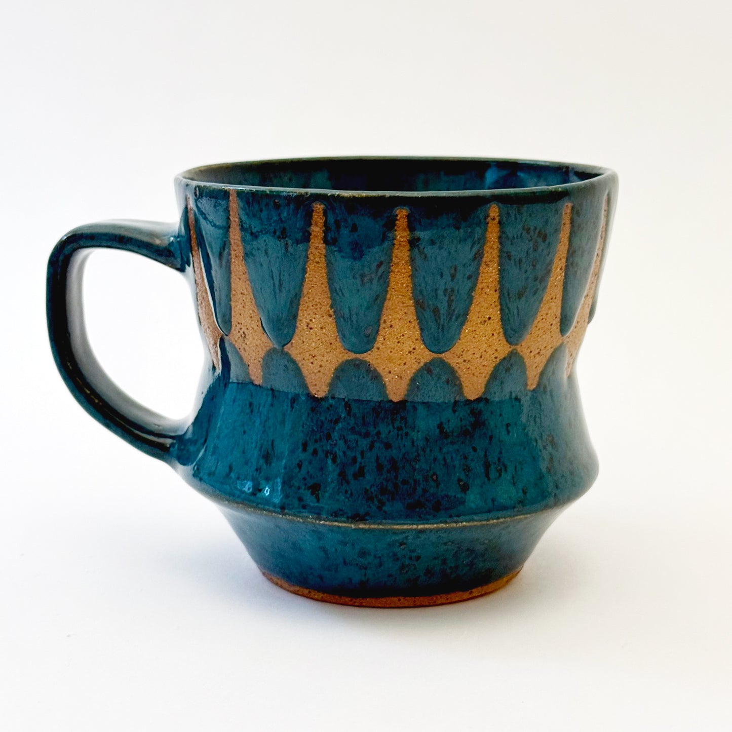 Hand-thrown stoneware mug with layered cobalt and teal blue glaze and a repeating retro geometric pattern in exposed speckled clay. The sculpted mug has a curved waist, angular handle, glossy finish, and mid-century-inspired design. Photographed against a soft white background.