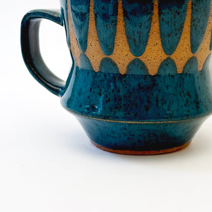 Hand-thrown stoneware mug with layered cobalt and teal blue glaze and a repeating retro geometric pattern in exposed speckled clay. The sculpted mug has a curved waist, angular handle, glossy finish, and mid-century-inspired design. Photographed against a soft white background.