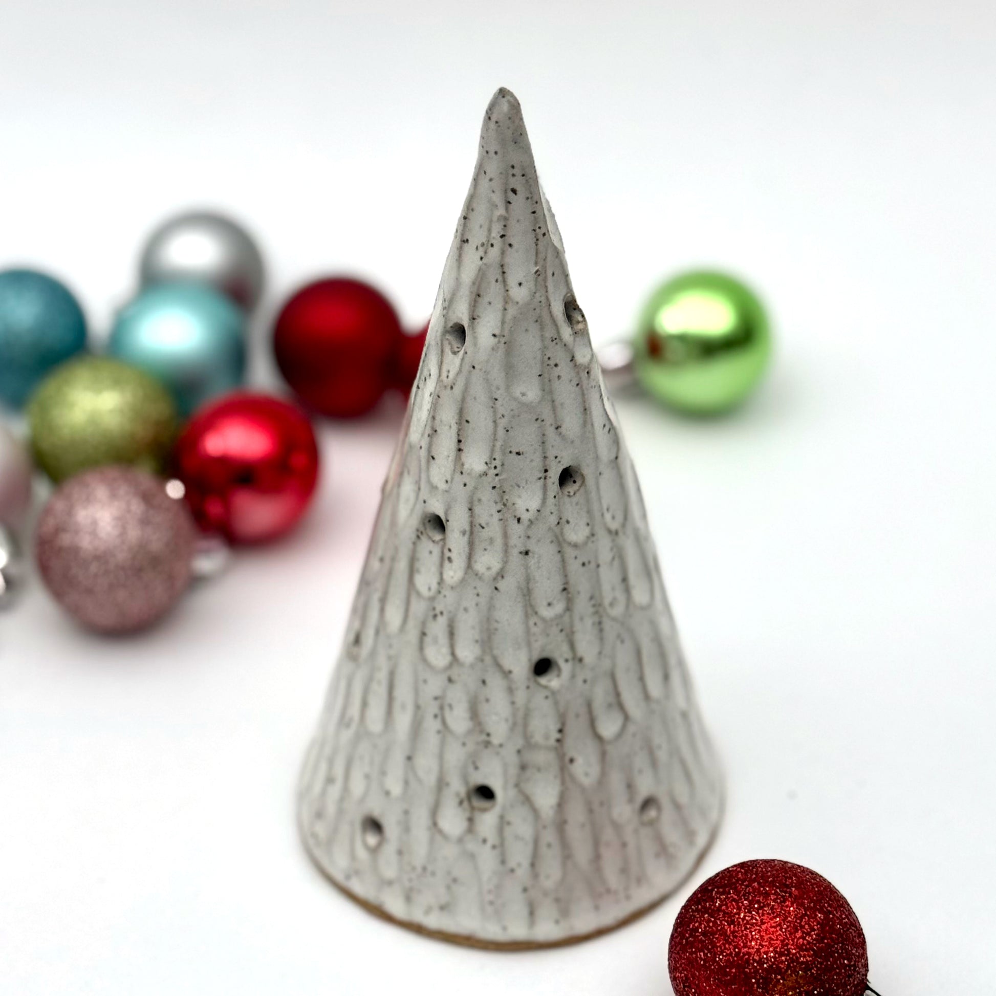 A textured white ceramic cone with multiple cutouts, resembling a glowing winter tree, displayed among colorful Christmas ornaments.