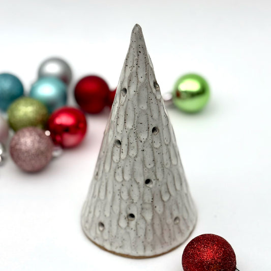 A textured white ceramic cone with multiple cutouts, resembling a glowing winter tree, displayed among colorful Christmas ornaments.
