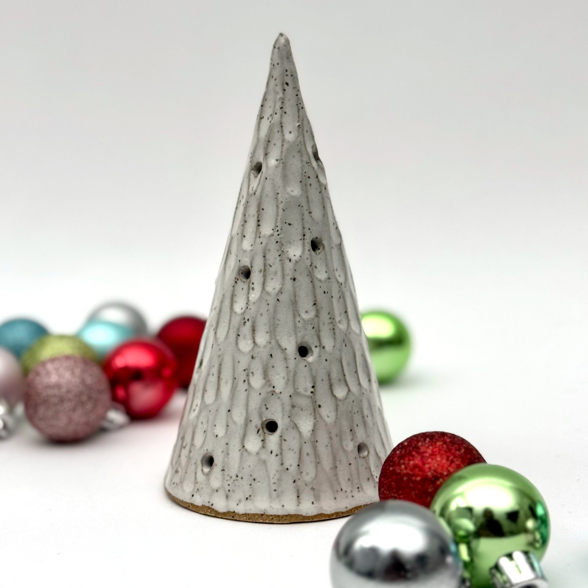 A textured white ceramic cone with multiple cutouts, resembling a glowing winter tree, displayed among colorful Christmas ornaments.