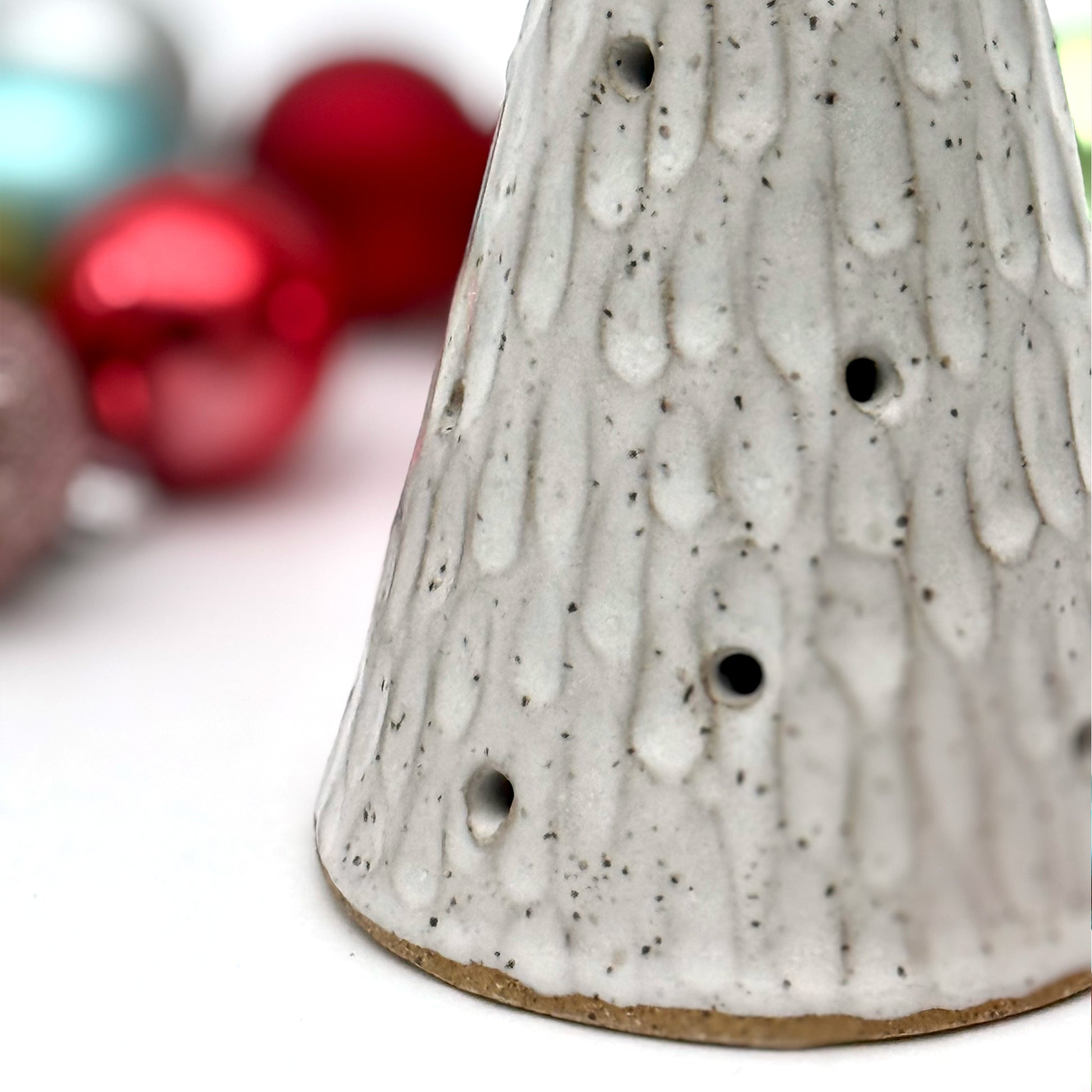 Close up of a textured white ceramic cone with multiple cutouts, resembling a glowing winter tree, displayed among colorful Christmas ornaments.