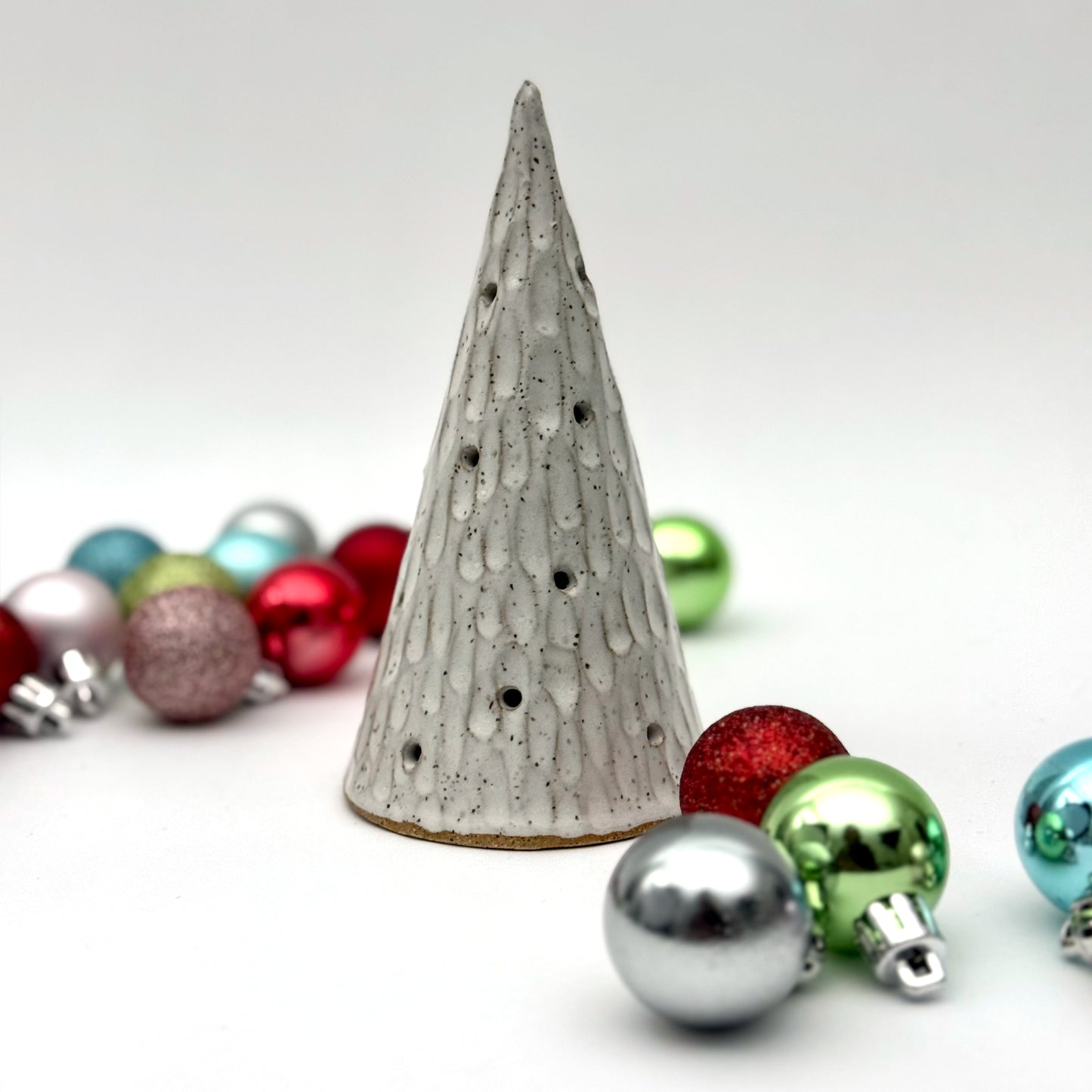 A textured white ceramic cone with multiple cutouts, resembling a glowing winter tree, displayed among colorful Christmas ornaments.