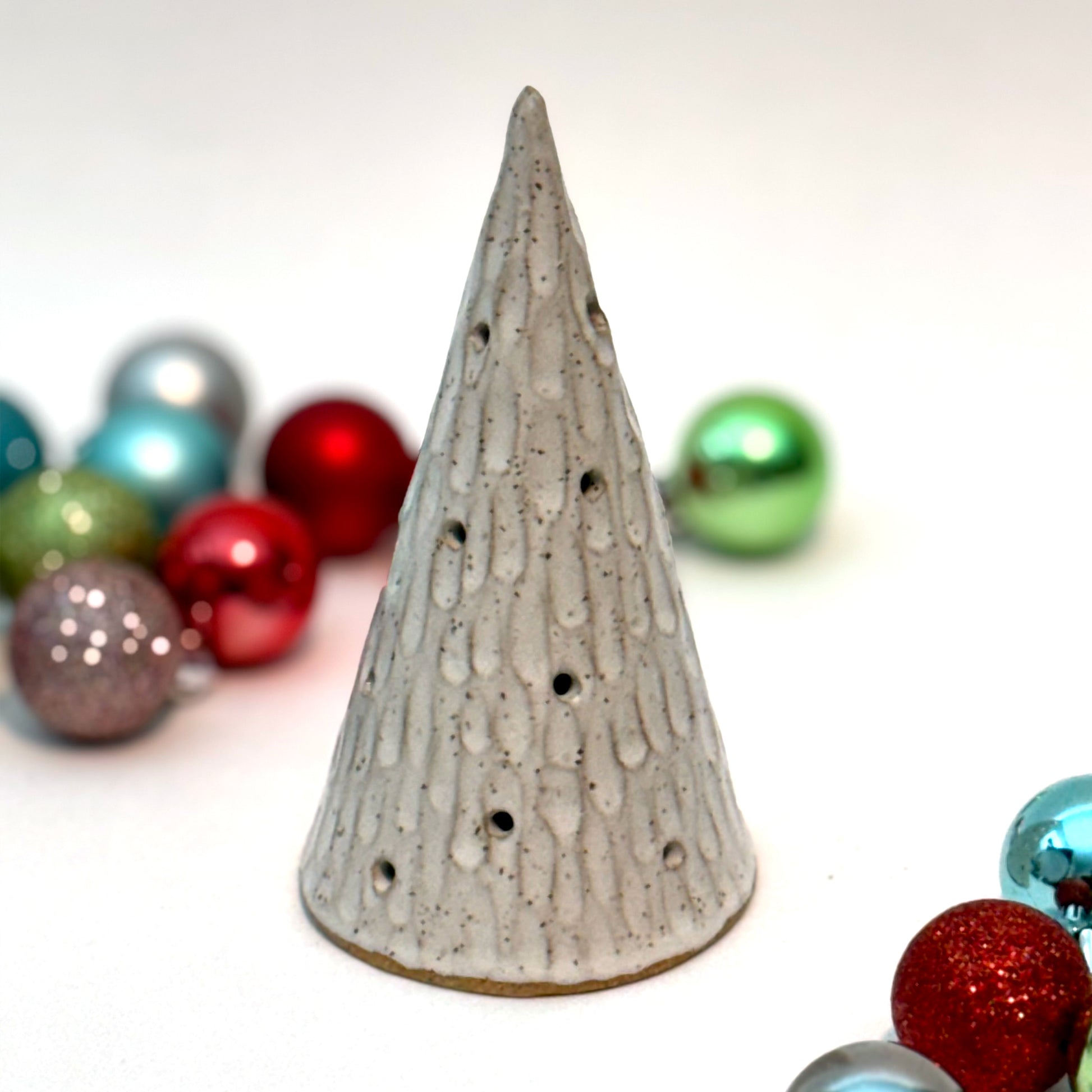 A textured white ceramic cone with multiple cutouts, resembling a glowing winter tree, displayed among colorful Christmas ornaments.