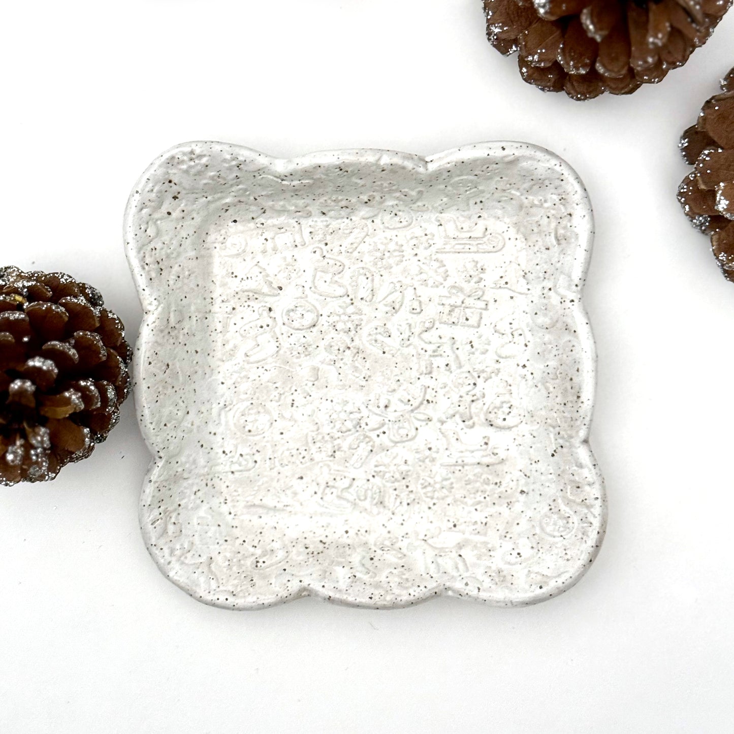 A square scalloped ceramic plate in speckled white with faint holiday embossing, surrounded by decorative pinecones.