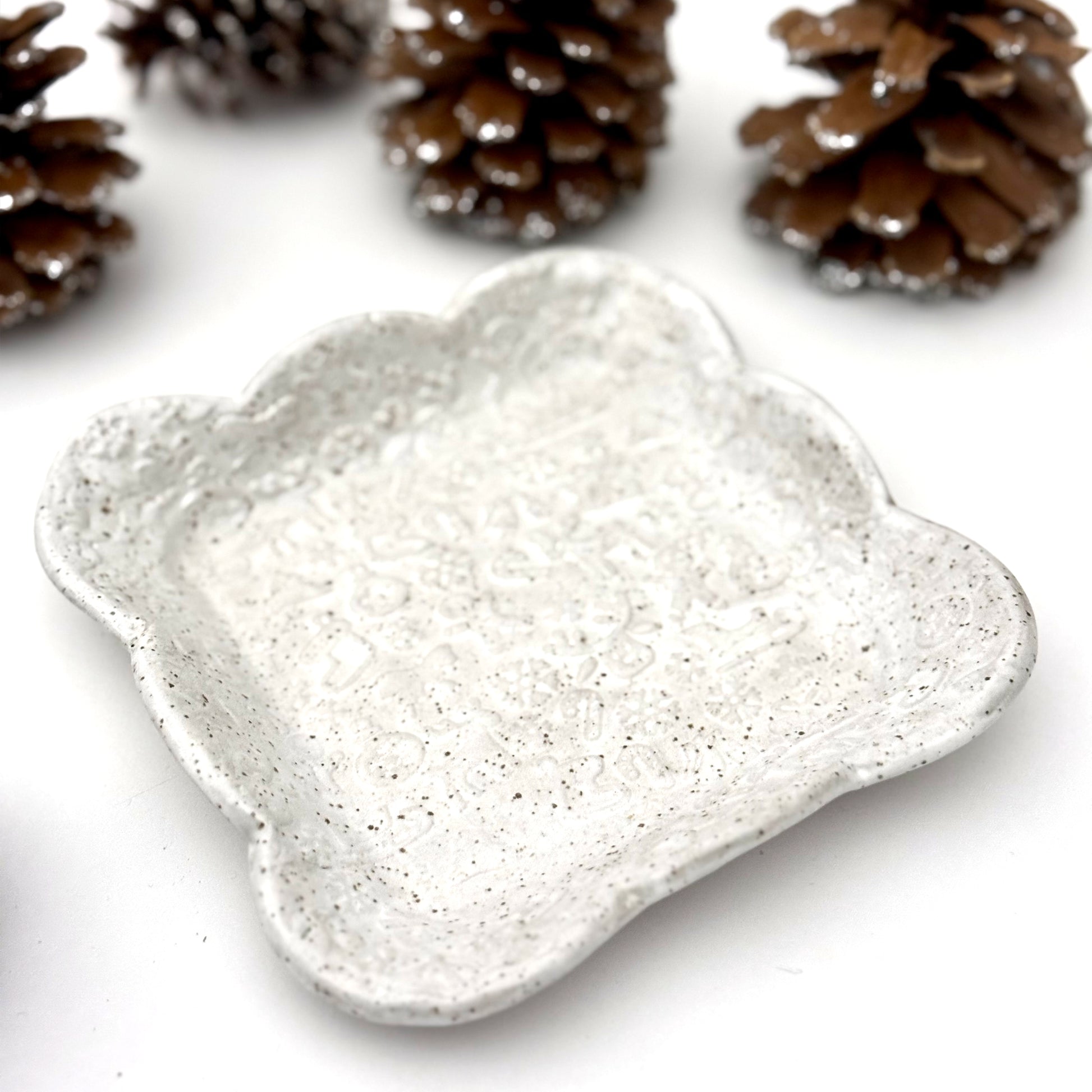 A square scalloped ceramic plate in speckled white with faint holiday embossing, surrounded by decorative pinecones.