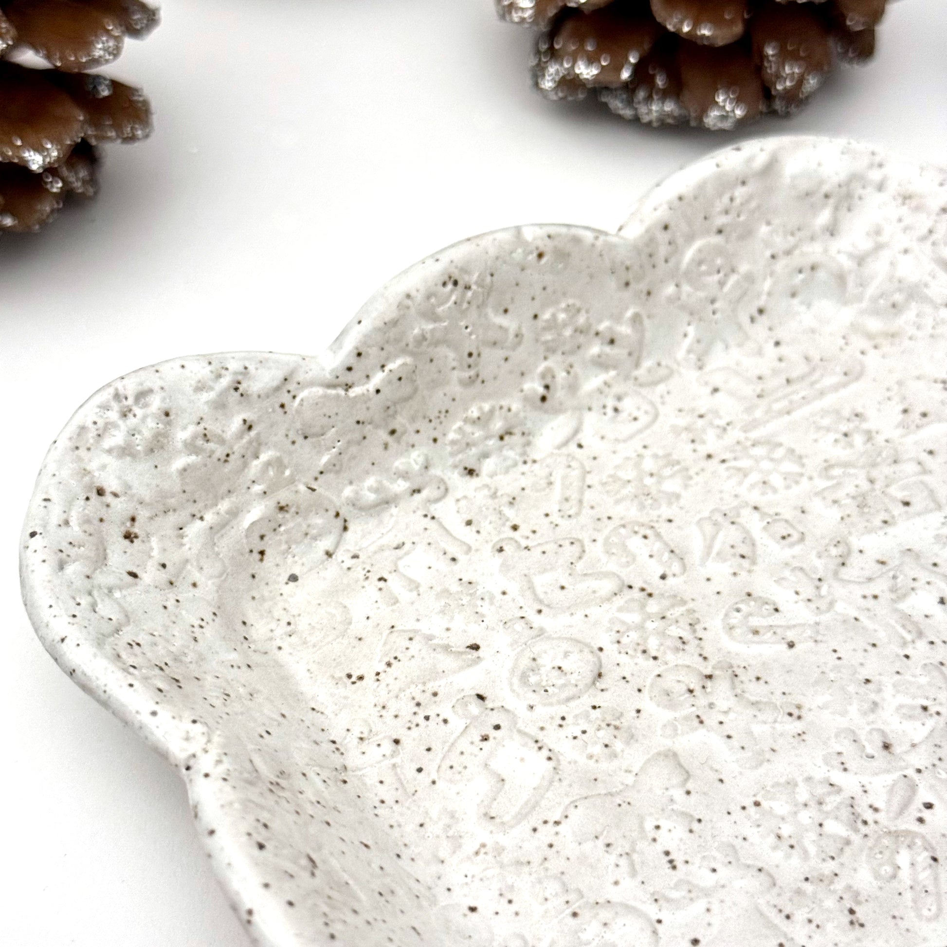 Close-up of a square scalloped ceramic plate in speckled white with faint holiday embossing, surrounded by decorative pinecones.