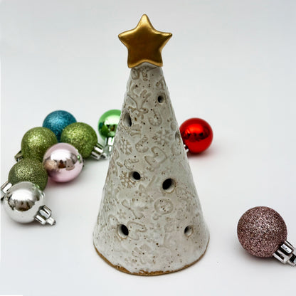 Ceramic Christmas tree luminary with gold star on top, surrounded by colorful miniature ornaments on a light background.