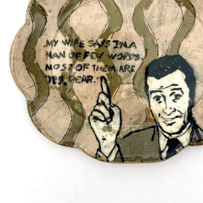 Close-up of a handmade ceramic trinket dish with a scalloped edge, featuring a vintage-style illustrated man pointing upward, wavy olive-green details, and a humorous handwritten quote.